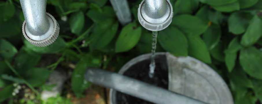 Das Foto zeigt einen rinnenden Wasserhahn. Das Wasser rinnt in eine Metallgießkanne.